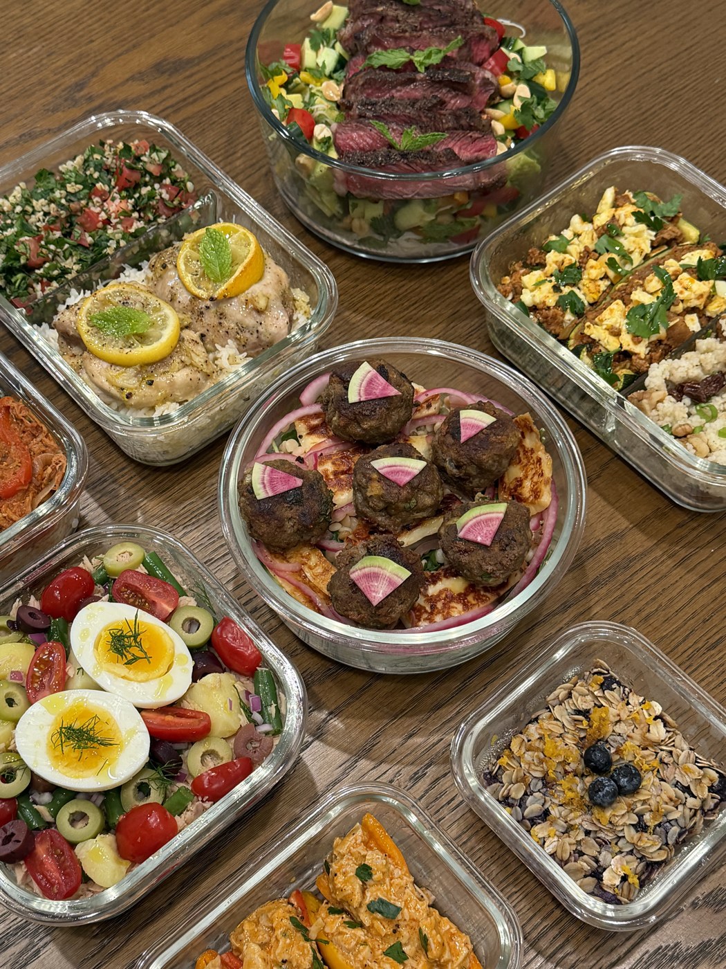 A spread of weekly meal prep — lamb meatballs over halloumi, seared steak salad, lemon chicken, tabbouleh, niçoise, and overnight oats